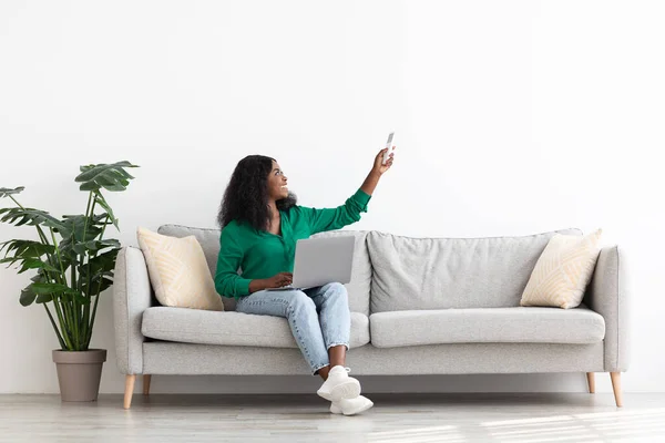 Glad svart kvinna sitter på soffan med laptop, håller fjärr — Stockfoto
