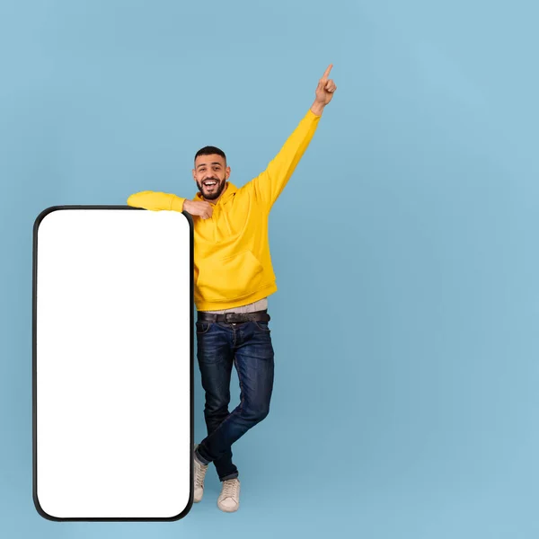 Aufgeregter arabischer Typ lehnt sich an Smartphone mit weißem leeren Bildschirm und zeigt nach oben, steht über blauem Hintergrund, Attrappe hoch — Stockfoto