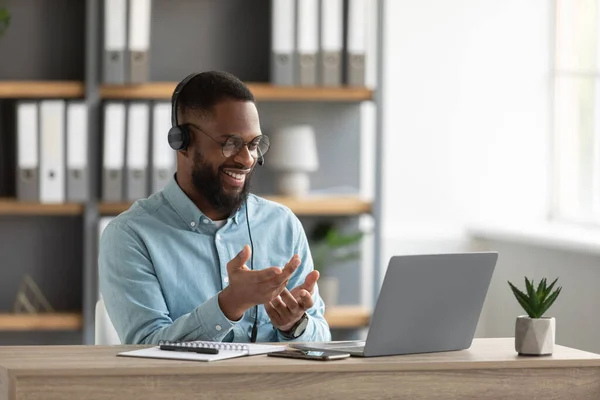 Happy millennial african bearded man in glasses and headphones gestures and works on laptop — стоковое фото