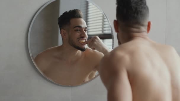 Hábitos saudáveis. Zoom em de jovem bonito muscular médio oriental homem escovar os dentes, olhando para o espelho no banheiro — Vídeo de Stock