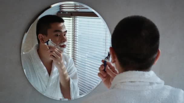 Японский мужчина бреется лицом, стоящим возле зеркала в ванной комнате — стоковое видео