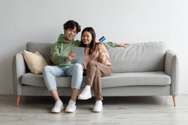 Kredi kartı ve tabletli genç Asyalı çift kanepede oturuyor, internetten mal sipariş ediyor, web mağazasından bir şeyler alıyor.
