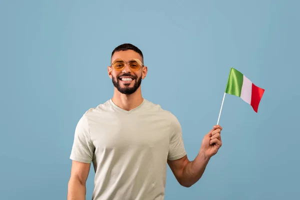 Pozitif Arap adam İtalya bayrağı gösteriyor, mavi arka planda poz veriyor ve kameraya gülümsüyor. Dil kursları konsepti — Stok fotoğraf