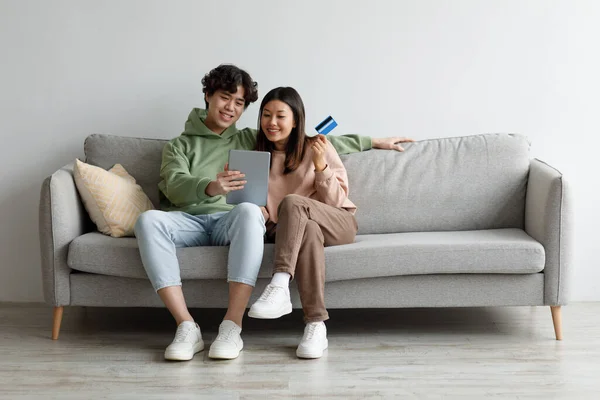 Jeune couple asiatique avec carte de crédit et tablette assis sur le canapé, commander des marchandises sur Internet, acheter des choses dans la boutique en ligne — Photo