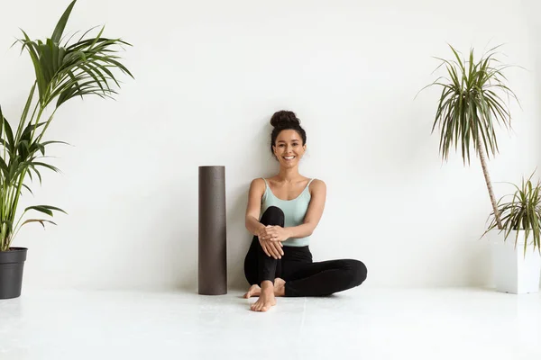 Concepto de bienestar. Hermosa hembra sonriente con estera de yoga sentada cerca de la pared blanca —  Fotos de Stock