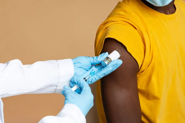 Felismerhetetlen orvos kék orvosi kesztyűt visel, hogy vakcinázza a fekete férfi beteget. — Stock Fotó