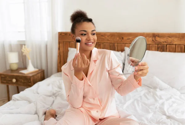 Vrolijke zwarte vrouw het aanbrengen van poeder of blusher met borstel, met behulp van decoratieve make-up in de voorkant van spiegel thuis — Stockfoto