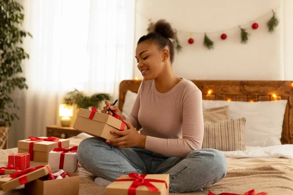Felicidades con las vacaciones de Año Nuevo. Feliz mujer negra escribiendo tarjeta de felicitación y envolviendo cajas de regalo, sentado en la cama — Foto de Stock