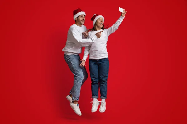 Hombre y mujer positivos usando sombreros de Papá Noel saltando y tomando selfie juntos — Foto de Stock