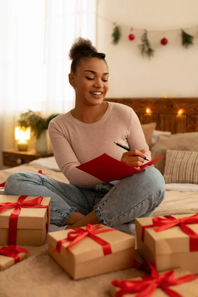 Excited black lady writing greeting card for friend, wrapping gift boxes and smiling, sitting on bed at home