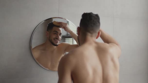 Над плечовим видом молодого сорочки без східного хлопця розчісує волосся, дивлячись на дзеркало у ванній кімнаті, збільшуючи знімок — стокове відео