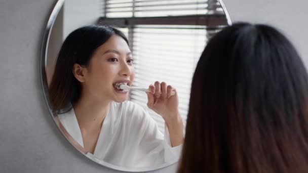 Concepto de rutina matutina. Hermosa mujer asiática cepillando dientes cerca del espejo en el baño — Vídeos de Stock