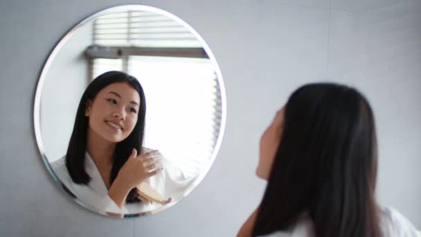 Mulher coreana feliz escovar o cabelo olhando para o espelho no banheiro — Vídeo de Stock