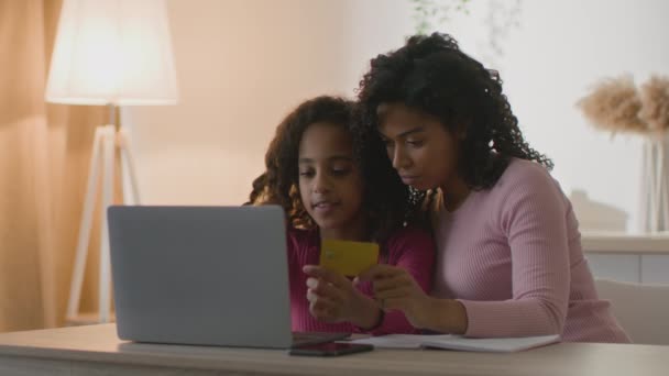Letramento financeiro. Mãe negra ensinando sua filha a comprar bens on-line, usando laptop e cartão de crédito em casa — Vídeo de Stock