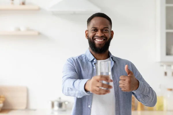 Sorridente ragazzo nero millenario mostra un bicchiere d'acqua in mano, pollice in alto in cucina interna — Foto Stock