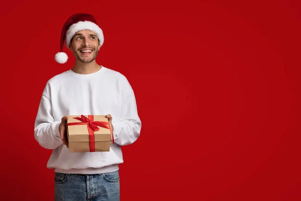 Verträumter junger Mann mit Weihnachtsmütze hält Geschenkbox und schaut weg — Stockfoto