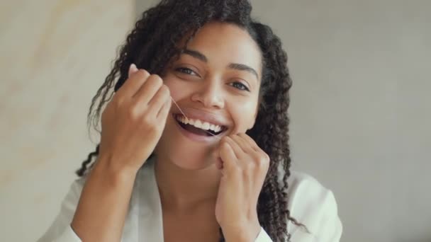 Зеркальный портрет молодой счастливой африканской женщины-американки, чистящей зубы зубной нитью в ванной утром — стоковое видео