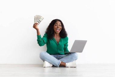 Rich african american lady gambling online, holding laptop and money