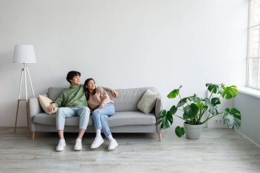 Spending weekend together. Happy millennial Asian couple resting on couch at home, empty space
