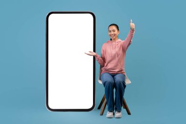 Joyful chinese woman sitting on chair, pointing at huge cellphone