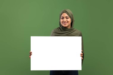 Happy muslim lady holding empty white placard for text advertisement, posing over dark green background, mockup