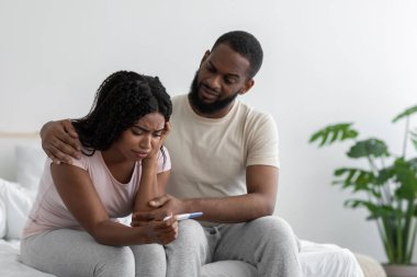 Unhappy young african american boyfriend calm sad girlfriend with pregnancy test, sit on bed
