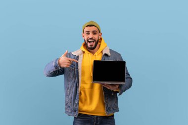 Overjoyed hipster arab guy pointing at laptop computer with blank screen, posing over blue studio background, mockup