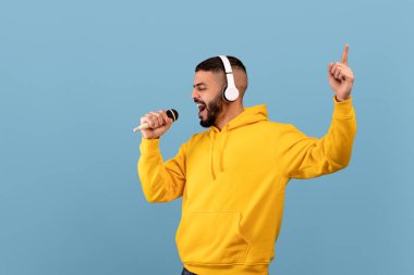 Joyful arab guy in headphones listening to music and singing song into microphone over blue studio background