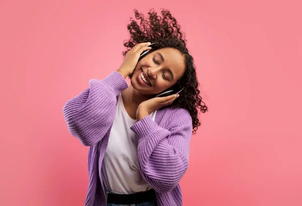 Carefree African American lady listening to music in wireless headphones, dancing with closed eyes on pink background — Stok fotoğraf