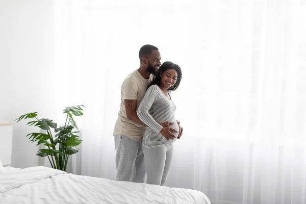 Happy millennial african american man in home clothes, hugs pregnant wife in white bedroom interior on window background — 图库照片