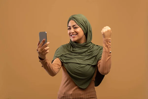 满意的穆斯林女性，头戴头巾，看着智能手机，表达喜悦，庆祝米色背景的成功 — 图库照片