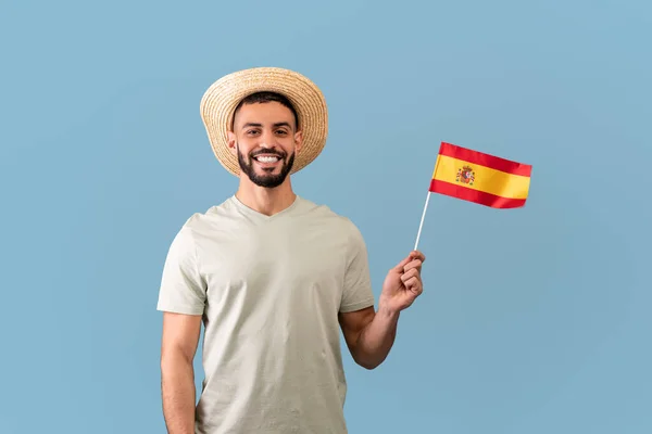 Happy arab man wearing t-shirt and hat, holding the spanish flag and smiling at camera, standing over blue background — Foto Stock