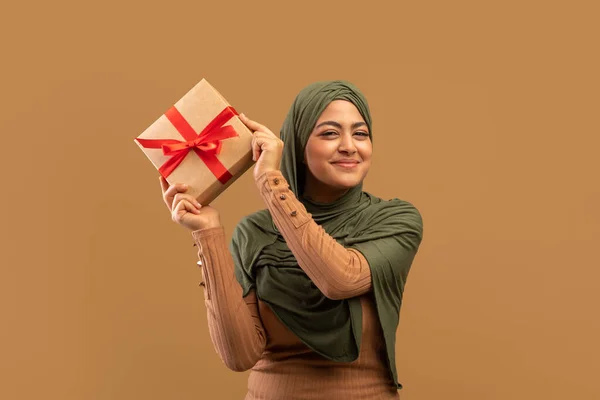 Happy muslim lady in hijab holding gift box, feeling excited to celebrate holiday, posing over beige studio background — 图库照片