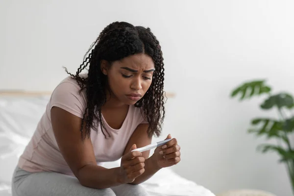 Unhappy sad young african american woman with pregnancy test sit on bed in white bedroom interior — Stockfoto