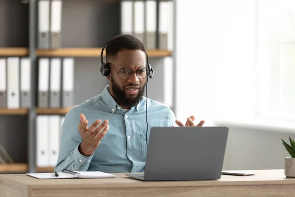 Descontento conmocionado triste joven afroamericano barbudo masculino en gafas, auriculares gestos y obras en PC — Foto de Stock