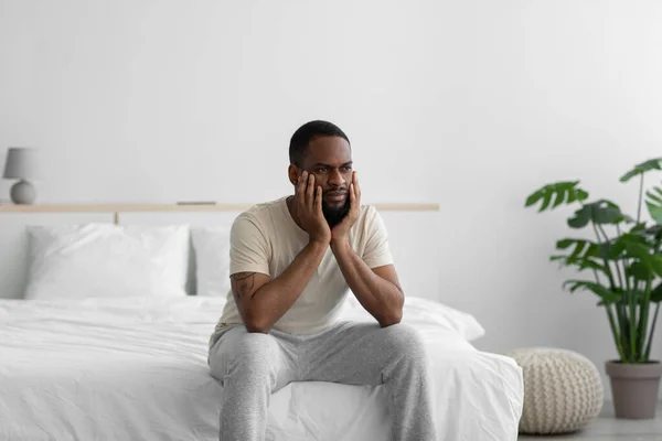 Θλιβερή φιλολογική χιλιετή αφροαμερικανός τύπος σε οικιακά ρούχα Ponders λύση στο πρόβλημα σε λευκό μινιμαλιστικό υπνοδωμάτιο — Φωτογραφία Αρχείου
