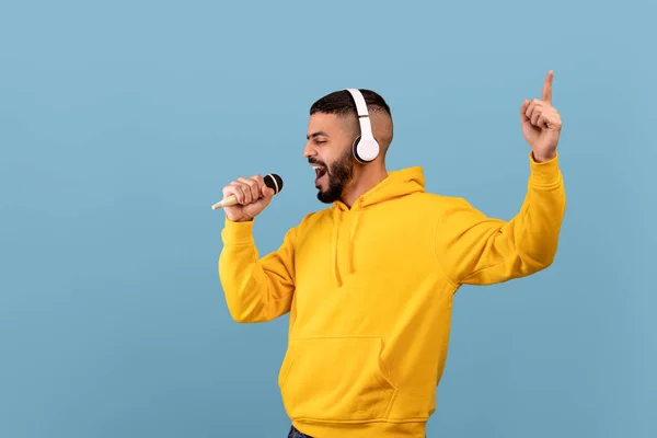 Alegre chico árabe en auriculares escuchando música y cantando canción en el micrófono sobre fondo de estudio azul — Foto de Stock