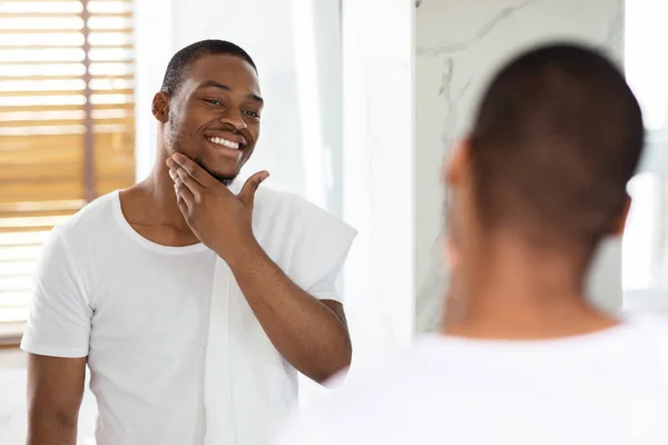 Cuidado de la piel masculina. guapo negro chico buscando en espejo en cuarto de baño, tocando barba — Foto de Stock
