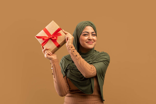 Happy muslim lady in hijab holding gift box, feeling excited to celebrate holiday, posing over beige studio background — Stock Image Happy muslim lady in hijab holding gift box, feeling excited to celebrate holiday, posing over beige studio background Stock Image
