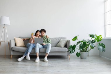 Asian couple using credit card and laptop, sitting on couch, ordering goods in web store, shopping online from home