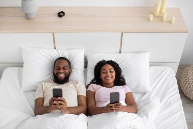 Smiling young african american husband and wife typing on phones, playing online games on bed in bedroom interior