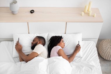 Sad young black guy and lady lie back to back, looks at smartphones on bed in bedroom interior