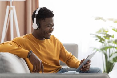 Happy black guy sitting on couch, watching movies on pad