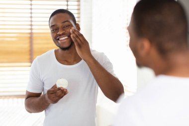 Male Skin Care. Handsome Black Guy Applying Moisturising Face Cream Near Mirror