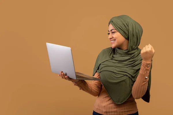 Νίκη στο διαδίκτυο. Ενθουσιασμένη μουσουλμάνα κυρία με χιτζάμπ κρατώντας φορητό υπολογιστή και γιορτάζοντας την επιτυχία με υψωμένη γροθιά, μπεζ φόντο — Φωτογραφία Αρχείου