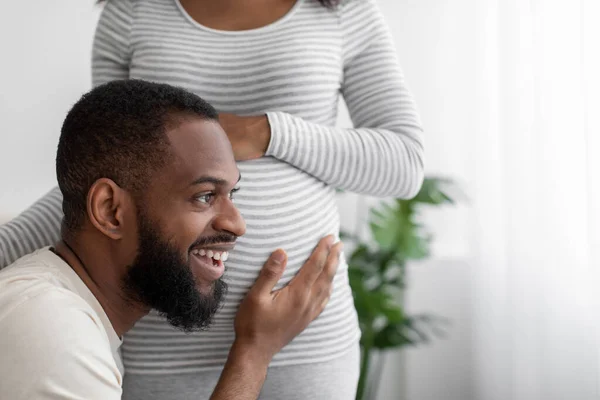 Glad happy millennial african american man listens to belly of pregnant wife in white bedroom interior, close up — стокове фото