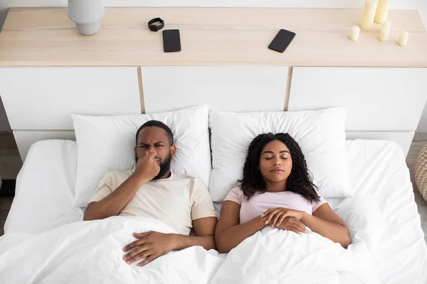 Sad disgruntled millennial african american husband closes nose from bad scent, wife sleeps on white bed together — Fotografie, imagine de stoc