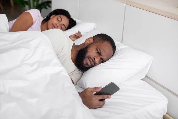 Unhappy millennial african american husband looks at phone, wife sleeps next to him on bed — Stok fotoğraf