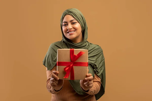 Cheerful arab lady in hijab holding gift box and smiling to camera, posing over beige studio background — Stock Photo, Image