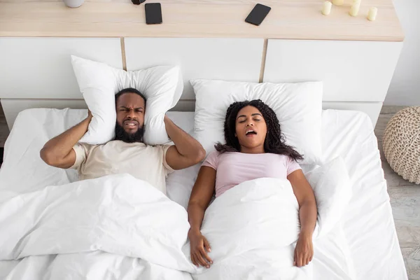 Angry disgruntled millennial black husband covers his ears with pillow, suffers from snoring and noise of wife at night — 스톡 사진